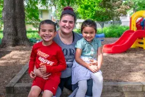 Photo of a white mother with her Latine daughter and son sitting on her lap at a playground